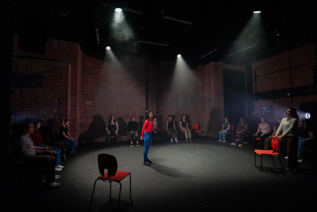 Action shot of the spring 2024 Shakespeare in Performance at RADA final presentation of Julius Caesar. Photo is taken from behind the audience, further away than others in this series. An actor wearing a bright red shirt stands with hands behind her back under a spotlight while another actor faces her, holding onto a chair. Other actors are seated behind and around them in mid-performance.